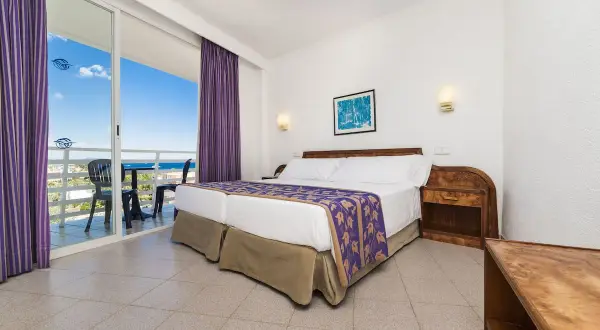 Pokój w hotelu Globales Palmanova Palace z widokiem na morze. Dwa łóżka, fioletowe zasłony i balkon.