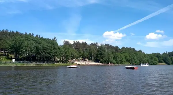 Letnie popołudnie nad jeziorem Osuszyno. Woda spokojna, na brzegu plażowicze, a na jeziorze łodzie. Może nawet jakieś ryby pływają pod powierzchnią.