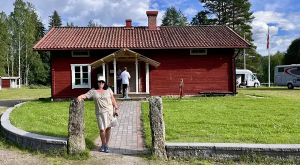 Värnäs camping: En kvinna står vid en röd stuga med tegeltak. Husbilar syns i bakgrunden under en blå himmel med moln.