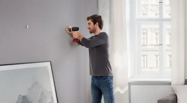 Uomo usa trapano per fissare listelli di legno al muro, preparando la parete per appendere un quadro.