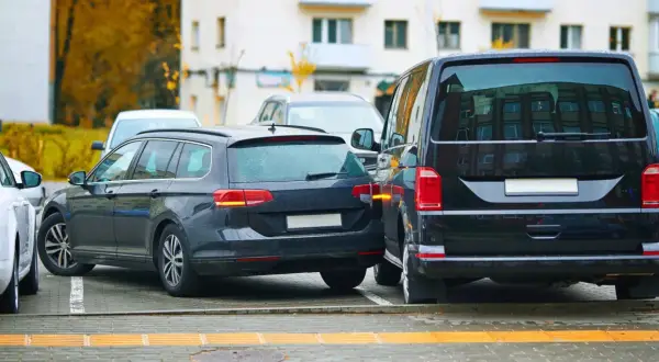 Ciemne kombi i van stoją blisko siebie na parkingu. Widać fragment białego auta. To strefa ruchu, gdzie parkowanie wymaga uwagi.