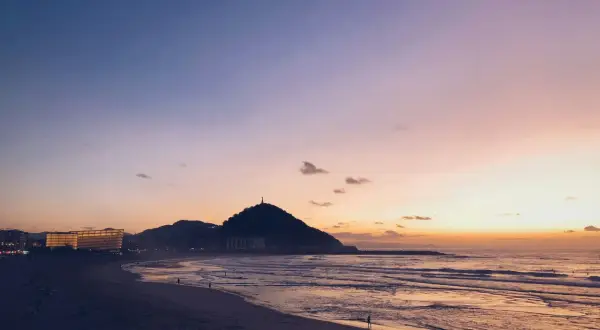 Atardecer en la costa San Sebastián. El cielo se tiñe de tonos rosados y naranjas sobre el mar y la playa, con la silueta del monte Urgull al fondo.