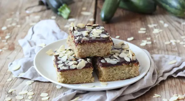 Saftiger Zucchini Kuchen mit Schokoladenglasur und Mandelblättchen, garniert mit frischen Zucchini im Hintergrund.