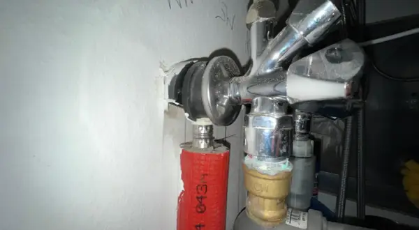 Chromglänzendes Eckventil der Wasserleitung mit rotem Schutzschlauch und sichtbaren Leitungen an einer weißen Wand.