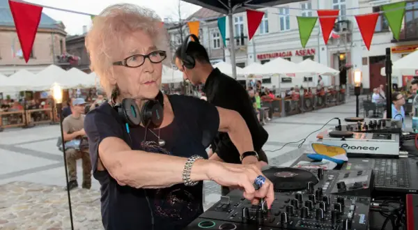 Starsza pani dj z pasją miksuje muzykę na placu miejskim, otoczona ludźmi i kolorowymi flagami.