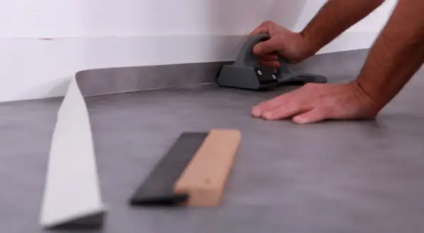 Pose d'un revêtement de sol souple, un rouleau gris est pressé contre le mur avec un outil.