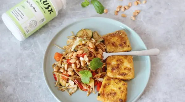 Tallrik med nudlar, grönsaker och friterad tofu. Bredvid en burk HUSK Naturlig fiber, perfekt för mat utan gluten och en balanserad mage.
