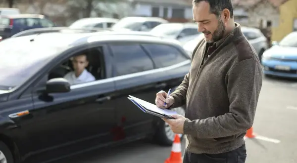 Examinador anotando en una carpeta. Un joven conductor espera en un coche negro. El examen práctico DGT y su precio son importantes para obtener el carnet.