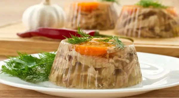 Schab po warszawsku w galarecie, ozdobiony marchewką i koperkiem, na talerzu. W tle czosnek i papryczka chili.