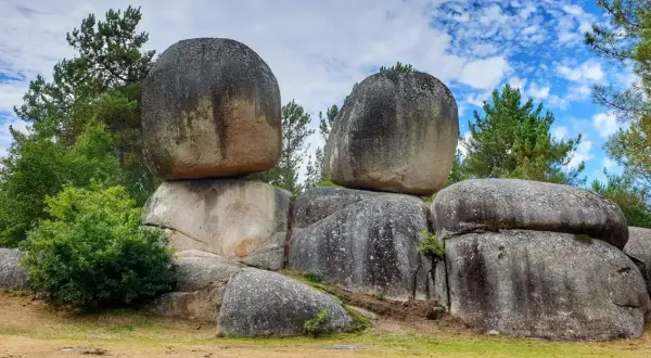 Gigantescas rocas apiladas, como penas de rodas, desafían la gravedad en un paisaje boscoso bajo un cielo azul con nubes.