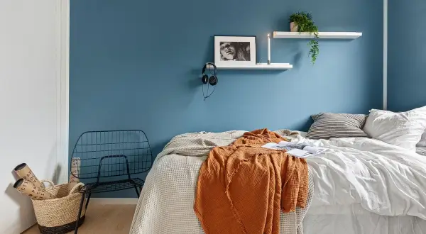 Camera da letto con pareti blu intenso, un letto accogliente con coperte arancioni e bianche, e una sedia in metallo nero. Ottime idee pareti bicolore per un ambiente moderno.