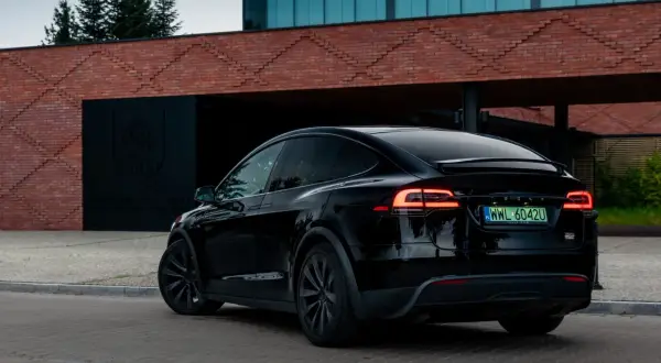 Czarna Tesla X z włączonymi światłami z tyłu, zaparkowana przed nowoczesnym budynkiem z cegły.