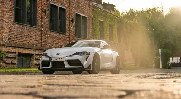 Biała Toyota GR Supra zaparkowana przed ceglanym budynkiem porośniętym bluszczem. Słońce przebija się przez drzewa, tworząc ciepłą, złotą poświatę.
