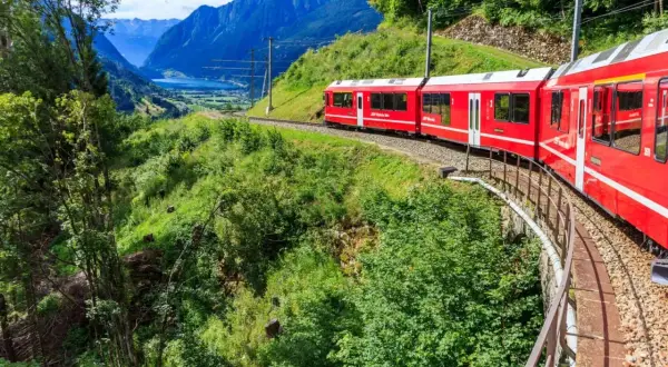 Röda tåget Bernina Express slingrar sig genom ett grönt alplandskap med en sjö och berg i bakgrunden. Undrar du över bernina express pris?