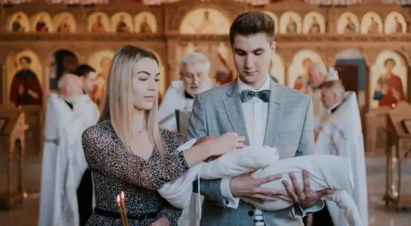 Młodzi rodzice trzymają dziecko do chrztu prawosławnego. W tle widać cerkiew i duchownych.