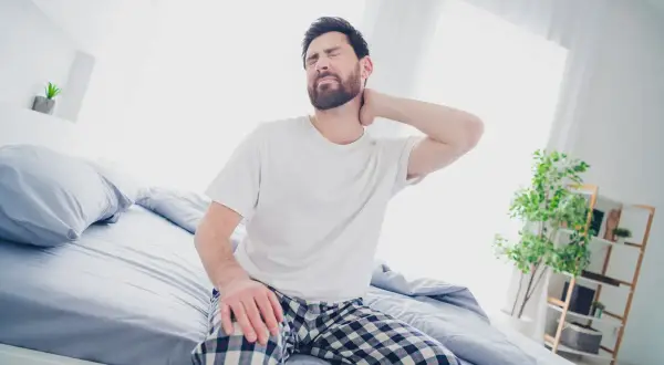 Uomo con torcicollo dopo aver dormito, si massaggia il collo con espressione sofferente, seduto sul letto.