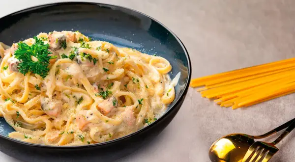 Delicioso plato de pasta con salmón y gambas, adornado con perejil fresco y queso rallado. Acompañado de espaguetis crudos y cubiertos dorados.
