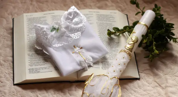 Biała szata, świeca z aniołkiem i gałązka symbol chrztu na otwartej Biblii.