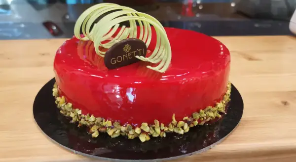 Torta con glassa a specchio lamponi, decorata con riccioli di cioccolato bianco e pistacchi tritati.