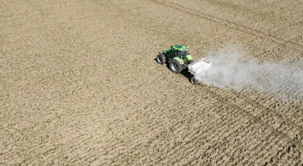 Zielony traktor z przyczepą rozsypuje nawóz na polu. To ważny etap prac polowych, który zapewnia prawidłowy wzrost roślin. Nikosar etykieta na przyczepie informuje o zawartości.