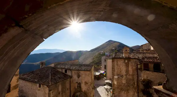 Solstrålar bryter fram genom en stenbåge över en pittoresk by i Abruzzo, Italien. Kulliga berg i bakgrunden.