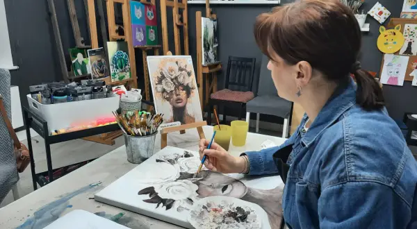 Kobieta w dżinsowej kurtce maluje obraz z kwiatami na płótnie w pracowni artystycznej.