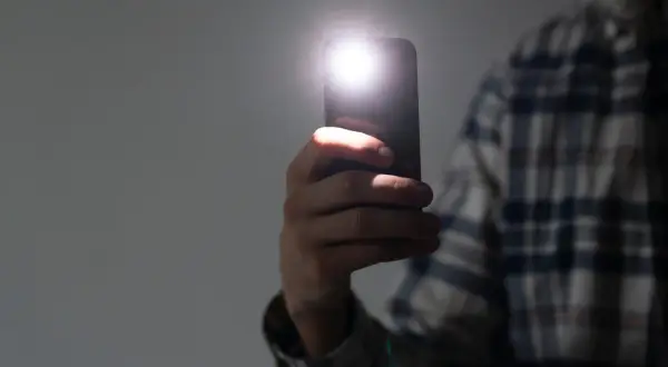 Dłoń trzyma telefon, którego lampa błyskowa rozświetla ciemność. Dowiedz się, jak włączyć lampę błyskową w telefonie, by zawsze mieć światło.