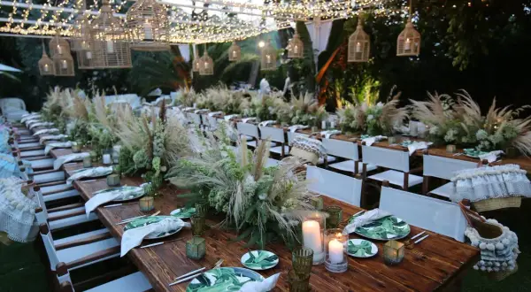 Tavoli in legno apparecchiati per un matrimonio in campagna, con centrotavola di fiori e pampas, luci soffuse e cesti decorativi.