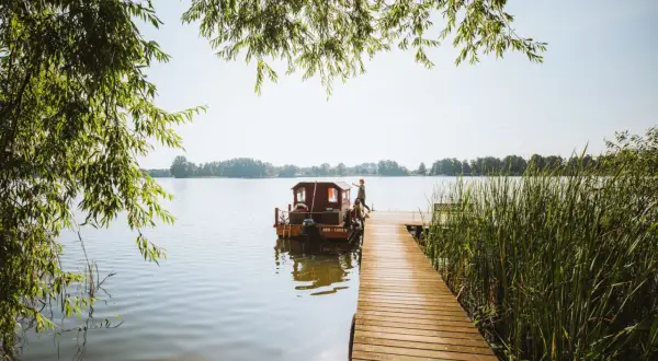 Ein Mann steht auf einem Holzsteg neben einem Hausboot auf einem See. Perfekter Urlaub in Brandenburg.