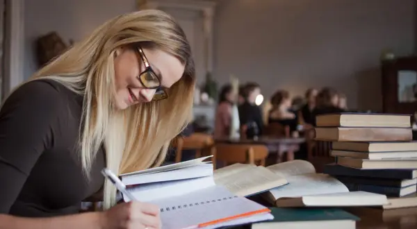 Kobieta w okularach pisze w notatniku, otoczona książkami. Marzy o studiach podyplomowych, na które czeka dofinansowanie.