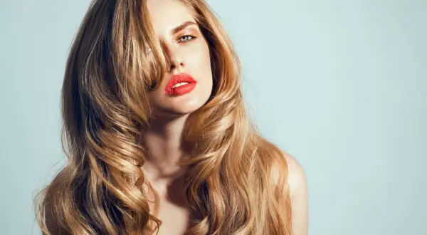 Kobieta z burzą falujących blond włosów, ułożonych w stylu butterfly cut, z wyrazistymi czerwonymi ustami.