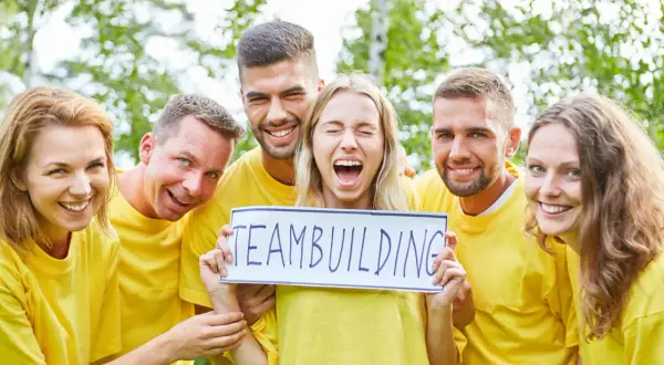En grupp glada människor i gula tröjor håller upp en skylt med "TEAMBUILDING". De verkar ha det roligt, som om de precis svarat på några snabba roliga frågor.