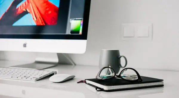 Na biurku iMac, klawiatura, myszka, notatnik z okularami i kubek. Higiena pracy to dbanie o porządek i komfort.