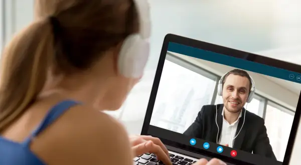 Kobieta w słuchawkach pisze na laptopie, prowadząc wideokonferencję z uśmiechniętym mężczyzną. On też ma słuchawki, ale ona pisze ale się nie umawia.