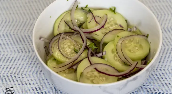 Deliciosa ensalada de pepino en rodajas finas con cebolla morada y hierbas frescas, servida en un cuenco blanco sobre un mantel azul y blanco.