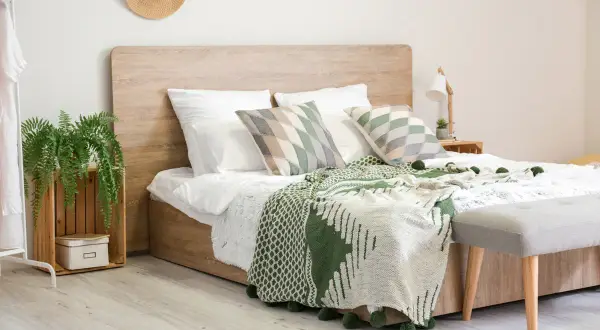 Gemütliches Schlafzimmer mit einem modernen **Aufbau Boxspringbett** in Holzoptik, grünen Decken und Kissen, sowie einer Hängepflanze.
