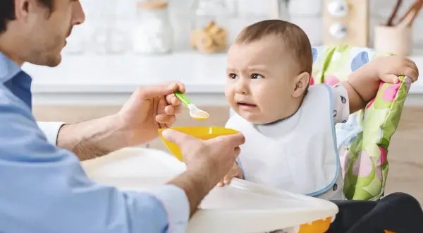 Ojciec karmi dziecko, które nie chce jeść. Czy niemowlę może jeść wątróbkę? Dziecko ma minę wyrażającą niechęć.