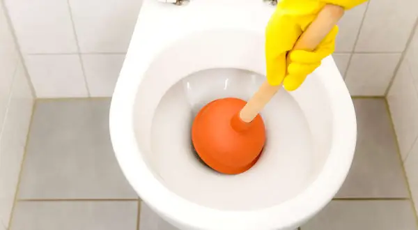 Mano con guanto giallo usa uno stantuffo arancione per liberare un filo d'acqua che scende nel water.