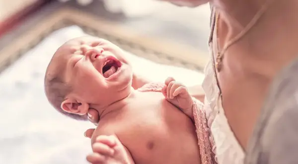 Bebé llorando con espasmos infantiles, envuelto en una manta rosa, sostenido por una persona.