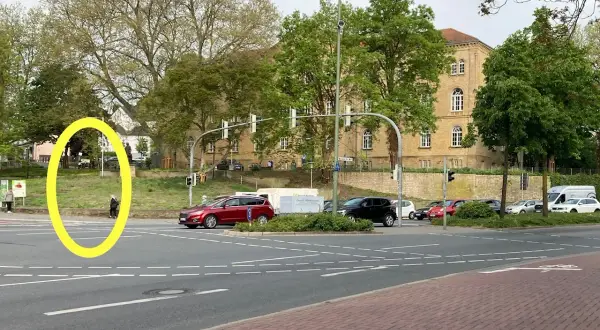 Eine Person wartet an der Ampel, um links abzubiegen. Autos stehen bereit, während im Hintergrund ein Gebäude und Bäume zu sehen sind.