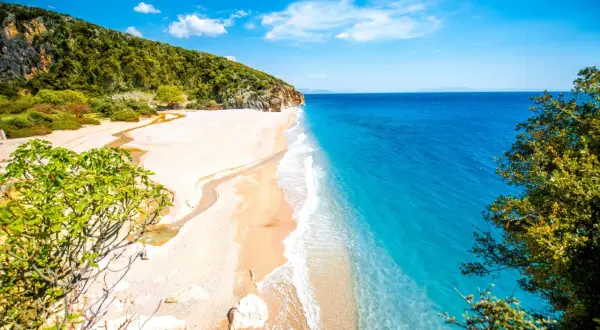 En orörd albanien strand med turkost vatten, vit sand och gröna kullar. En liten flod rinner ut i havet.