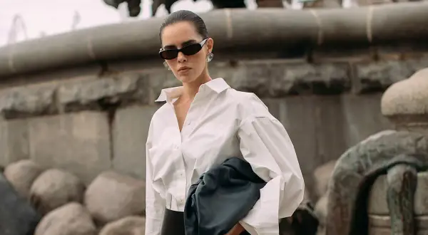 Elegancka kobieta w białej koszuli i okularach przeciwsłonecznych, z czarną torebką. Stylizacja godna polskiego domu mody.