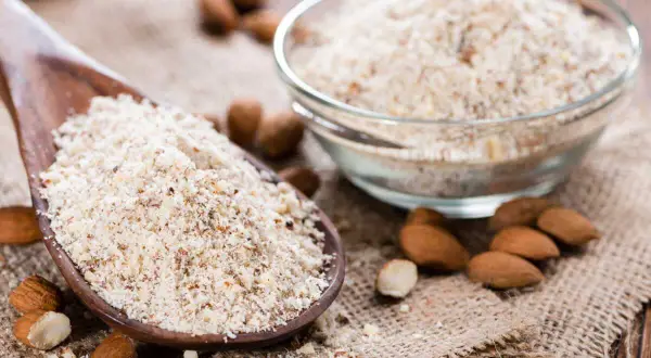 Cuillère en bois remplie de poudre d'amande, idéale pour remplacer la farine dans vos recettes. Amandes entières et bol de poudre à côté.