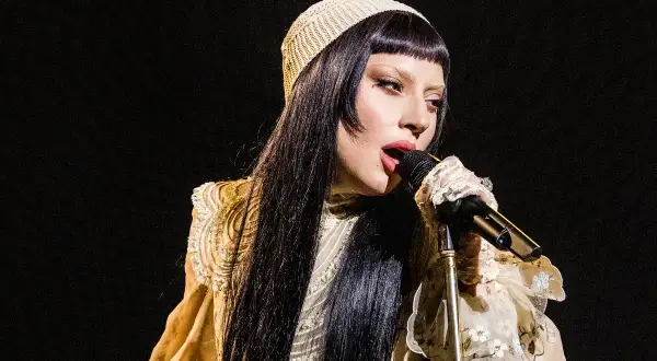 Lady Gaga w beżowym nakryciu głowy i ozdobnej sukni śpiewa do mikrofonu.