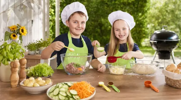 Zwei Kinder mit Kochmützen bereiten sich auf das Grillen vor. Sie mischen Zutaten für Burger und haben Spaß beim gemeinsamen Kochen.