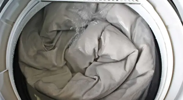 A grey comforter is being washed in a washing machine, showing the best way to wash a comforter. Water is splashing onto the fabric.