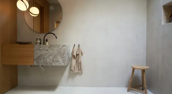 Modernt badrum med handfat i marmor och väggar i microcement. En rund spegel och två lampor ovanför.