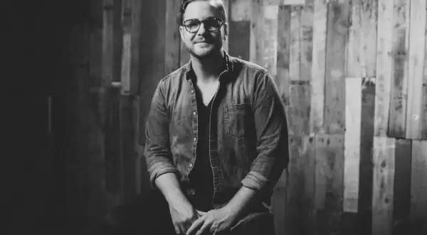 Jordan Reynolds, a talented songwriter, poses confidently in a denim shirt against a rustic wooden backdrop.