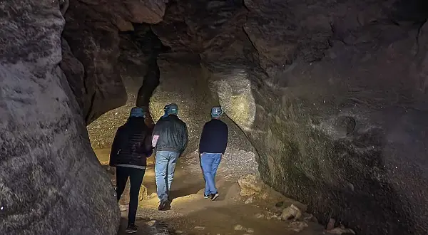 Tres personas con cascos exploran una cueva oscura, sus linternas iluminan las paredes rocosas y el suelo irregular. Los yesos naturales brillan en la oscuridad.