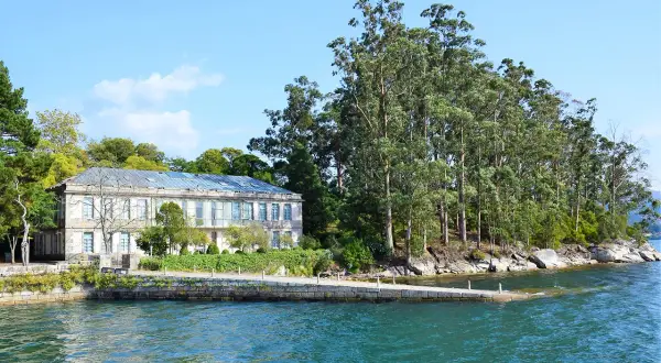 Edificio histórico en la illa de San Simón, rodeado de exuberante vegetación y aguas turquesas.
